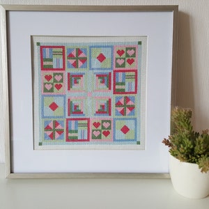 Op de afbeelding: Een ingelijst kruissteekontwerp met een patchwork-patroon in verschillende kleuren, waaronder rood, roze, groen en blauw. De quilt is gemaakt van vierkanten met verschillende ontwerpen, waaronder harten, sterren en geometrische vormen.
