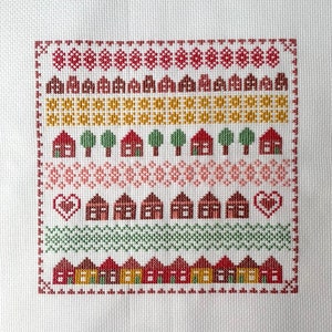 May include: Cross-stitch pattern featuring a row of red houses with yellow windows and green trees, surrounded by a border of red, pink, and yellow geometric shapes.