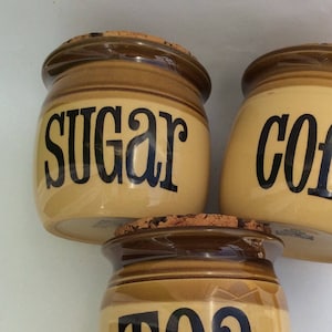 Può includere: Tre barattoli in ceramica con coperchi in sughero. I barattoli sono di colore marrone e beige con scritte nere. I barattoli sono etichettati con "Zucchero", "Caffè" e "Tè".