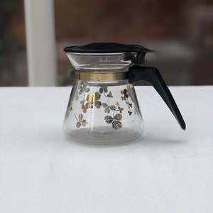 Peut inclure: Une cafetière en verre transparent avec une poignée et un couvercle noirs. La cafetière présente une bande dorée et un motif de motifs en forme de trèfle dorés et argentés. La cafetière est posée sur une surface blanche.