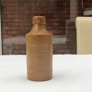 Puede incluir: Una botella de cerámica marrón con la parte superior redondeada y una etiqueta que dice "STAFFORDSHIRE POTTERY" en el lateral.