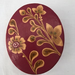 May include: An oval trinket box with a burgundy base and a floral design painted in gold and green.