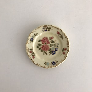 Op de afbeelding: Een klein, wit keramiek bord met een gouden rand en een bloemenontwerp. De bloemen zijn roze, blauw en groen.