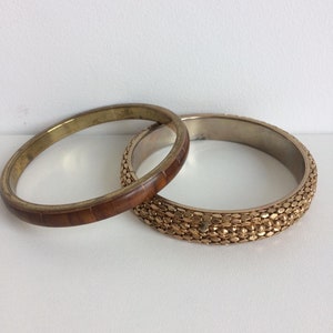 May include: Two bangles. One is gold-toned with a brown, textured band. The other is a gold-toned bangle with a textured, scale-like pattern. Both are set against a white background.
