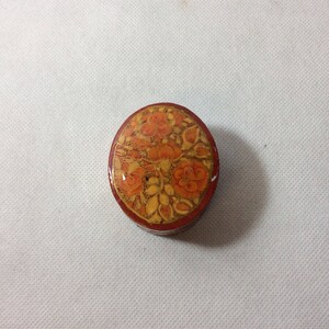 May include: A round, wooden trinket box with a red rim and a floral design painted in orange and yellow.