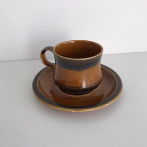 Puede incluir: Un juego de taza y platillo de café de cerámica con rayas marrones y negras. La taza tiene una forma redondeada y un asa. El platillo es redondo y tiene un borde ligeramente elevado.