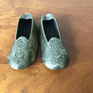 Può includere: Un paio di piccole scarpe antiche in ottone con motivi floreali. Le scarpe presentano disegni incisi di fiori e foglie. Le scarpe sono esposte su una superficie di legno.
