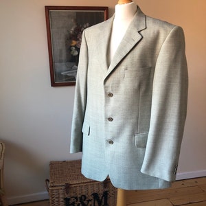 Puede incluir: Una chaqueta de sport gris claro con cierre de tres botones. La chaqueta está en un maniquí y tiene un patrón sutil.