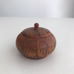 May include: A small, round wooden box with a lid. The box is made of dark wood with a natural grain pattern. The lid has a small knob on top.