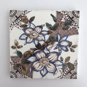 May include: A white ceramic tile with a floral design. The tile features three white flowers with blue outlines and brown centres. The flowers are surrounded by green leaves and a brown geometric pattern.