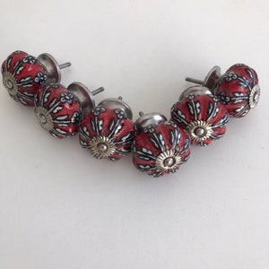 Pode incluir: Seis botões de armário de cerâmica vermelha e preta com detalhes prateados. Os botões têm um padrão floral e estão dispostos em semicírculo.