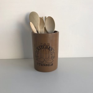 Puede incluir: Porta utensilios de cocina de cerámica marrón con las palabras "Kitchen Utensils" y un gráfico de tres utensilios. El porta utensilios está lleno de cucharas de madera.
