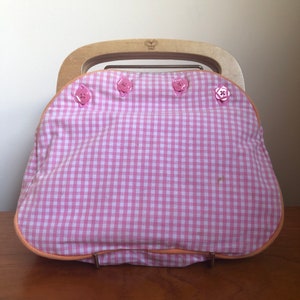 May include: A pink and white gingham fabric handbag with a wooden handle and pink buttons. The bag has a zipper closure and a butterfly logo on the handle.