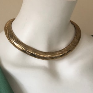 Puede incluir: Un collar de gargantilla dorado y acanalado exhibido en un maniquí blanco. El collar tiene un diseño grueso y redondeado y una superficie texturizada. La joya es una pieza llamativa, perfecta para añadir un toque de glamour vintage a cualquier atuendo.