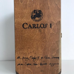 Op de afbeelding: Een houten doos met een zwart-wit logo met de tekst "Carlos I". De doos heeft een handgeschreven inscriptie met de tekst "El brandy Carlos I de Pedro Domecq es un Solera Gran Reserva envejecido".
