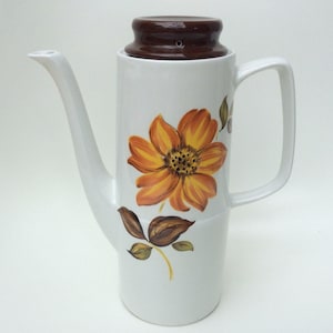Może przedstawiać: Biały ceramiczny dzbanek do kawy z brązową pokrywką i kwiatowym wzorem z dużym pomarańczowym kwiatem i brązowymi liśćmi.