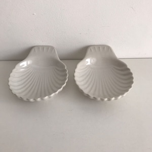 May include: Two white ceramic seashell-shaped dishes. The dishes have a ribbed texture and are perfect for serving small snacks or condiments.