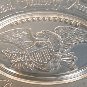 May include: A clear glass plate with an etched design of the United States of America seal. The seal features a bald eagle with outstretched wings, a shield with vertical stripes, and an olive branch and arrows.