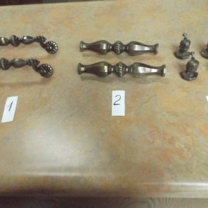 May include: Three sets of antique brass cabinet hardware. The first set is two ornate pulls, the second set is two long pulls, and the third set is four small knobs.
