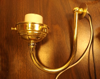 May include: A gold-toned metal wall sconce with a curved arm and a white porcelain socket. The sconce has a round base and is attached to a wall with screws.