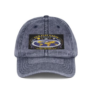 May include: A navy blue acid-washed baseball cap featuring a rectangular patch on the front. The patch displays a graphic of a flat earth map with yellow continents, blue oceans, and the text "OUR FLAT EARTH THE GREATEST SECRET OF THIS AGE".
