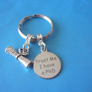 May include: Silver keychain with a graduation cap charm and a round tag that says "Trust Me I have a PhD".