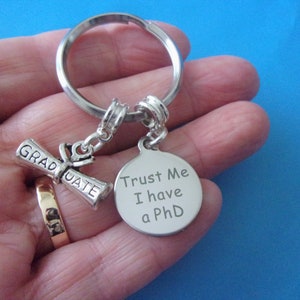 May include: Silver keychain with a graduation cap charm and a round tag that says "Trust Me I have a PhD".