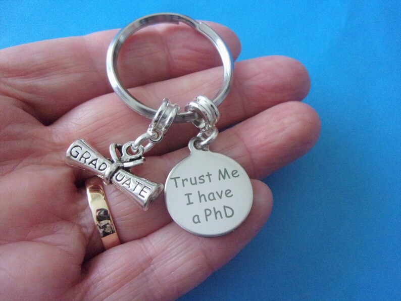 May include: Silver keychain with a graduation cap charm and a round tag that says "Trust Me I have a PhD".