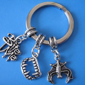 May include: Silver-coloured keyring with three charms: a stylized bat, a set of vampire teeth, and the words "bite me" in cursive. The keyring is on a blue background.