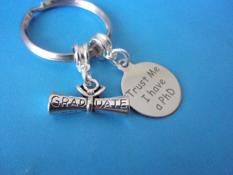 May include: Silver keychain with a graduation scroll charm and a round tag that says "Trust Me I have a PhD".