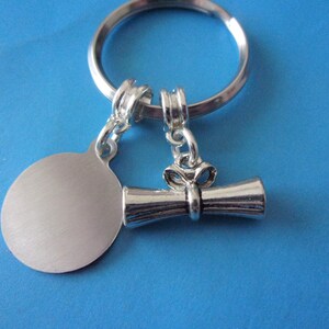 May include: Silver key chain with a round tag and a graduation cap charm. The tag is blank and the charm is a scroll with a bow.