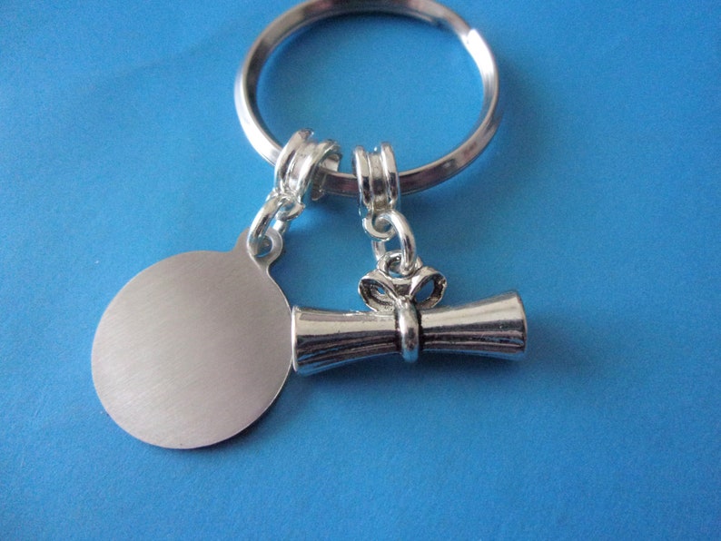 May include: Silver key chain with a round tag and a graduation cap charm. The tag is blank and the charm is a scroll with a bow.