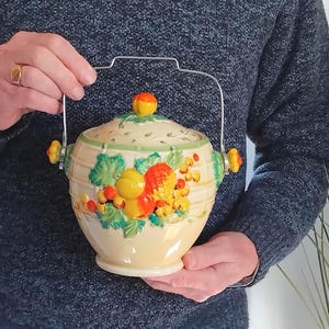 May include: A cream-coloured ceramic jar with a lid and a metal swing handle. The jar features a raised, colourful relief design of peaches, grapes, and green leaves on the front. The lid has a small orange knob, and the handle is silver-toned.