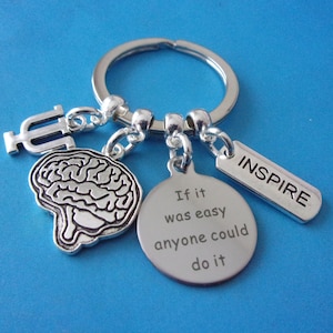 May include: Silver keyring with a brain charm, a charm with the text "If it was easy anyone could do it", a charm with the text "INSPIRE", and a charm with the Greek letter Psi.