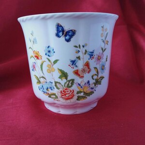 May include: White ceramic planter decorated with flowers and butterflies. The design includes colourful flowers and two butterflies in blue and orange. The planter has a scalloped base and a decorative rim. The planter is suitable for indoor use.