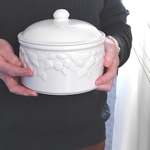 May include: A white ceramic canister with a matching lid, featuring a raised, embossed fruit and leaf pattern around the upper circumference of the cylindrical body. The container has a smooth, glossy finish and a simple knob handle on the lid.