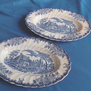 May include: Two oval-shaped ceramic platters featuring a white base with a blue transferware pattern. The design depicts a rural cottage scene in the centre, surrounded by a detailed floral and leaf border with a scalloped, fluted edge.