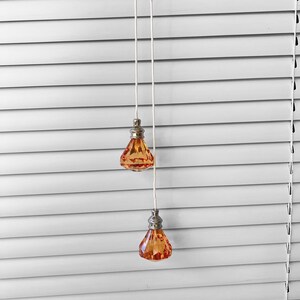 May include: Two orange crystal pull cords hanging from white blinds. The cords have a silver metal top.