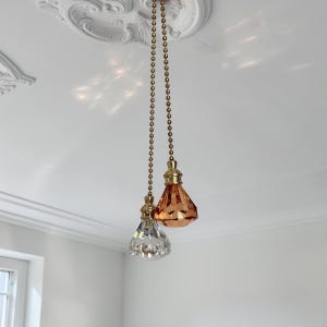 May include: Two ceiling fan pull chains featuring faceted, diamond-shaped finials. One finial is clear glass, and the other is amber-colored glass. Both are attached to gold-toned metal ball chains and hardware, hanging from a white ceiling.
