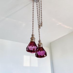 Könnte beinhalten: Zwei Deckenventilator-Zugketten mit facettierten, rautenförmigen violetten Glasanhängern. Jeder Anhänger ist an einem silberfarbenen Metallverbinder und einer langen, silberfarbenen Kugelkette befestigt, um Deckenventilatoren oder Leuchten zu bedienen.