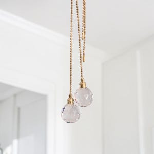 Op de afbeelding: Twee trekkoorden voor plafondventilatoren met goudkleurige metalen kogelkettingen en gefacetteerde, heldere glazen bolvormige ornamenten. Elk ornament is met een goudkleurige metalen dop aan de ketting bevestigd als decoratief accessoire.