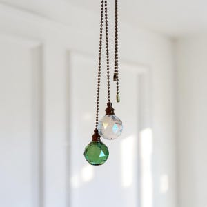 May include: Two ceiling fan pull chains featuring faceted spherical ornaments. One ornament is clear glass and the other is emerald green glass. Both are attached to dark bronze-toned metal ball chains with matching dark bronze-toned connectors.