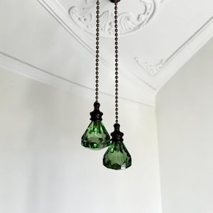 Könnte beinhalten: Zwei grüne, facettierte Glas-Lichtzugschalter an langen, bronzefarbenen Metallkugelketten. Jeder Zugschalter besteht aus einem rautenförmigen Glasanhänger mit einer dunklen Metallkappe an der Oberseite zur Bedienung von Deckenventilatoren.