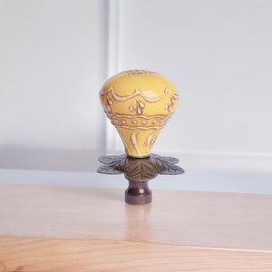 May include: A decorative yellow bottle stopper with a floral design. The stopper has a bulbous top with intricate detailing and a dark brown base with a leaf-shaped design. The stopper is made of ceramic and metal.