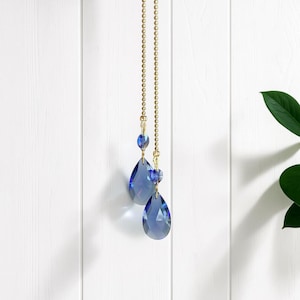 Puede incluir: Un adorno colgante decorativo que presenta dos cristales azules facetados en forma de lágrima suspendidos de una cadena de bolas dorada. Cada cristal está acentuado con una cuenta azul redonda más pequeña en la parte superior.
