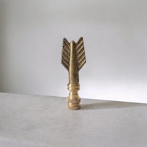 May include: A gold metal lamp finial with an arrowhead design. The finial is about 2 inches tall and has a slightly worn finish.