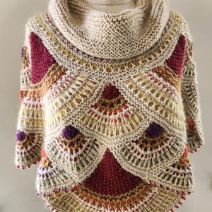 以下が含まれることがあります： ベージュの手編みポンチョ、ハイネック。ポンチョは、バーガンディ、ゴールド、紫色の幾何学模様が特徴です。裾には小さな赤いビーズが飾られています。マネキンに着せられています。