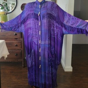 Op de afbeelding: Een lange paarse en blauwe robe met een witte print van figuren en franjes. De robe heeft een knoopsluiting aan de voorkant en twee zakken.