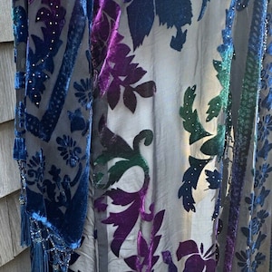 Peut inclure: Une robe noire transparente avec un motif floral dans les tons de bleu, vert et violet. La robe est ornée de perles irisées et possède une bordure à franges.