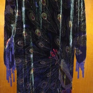 Peut inclure: Longue robe en velours bleu foncé avec un motif de plumes de paon. La robe présente des accents dorés et rouges, avec des franges aux manches et à l'ourlet. Le design général est élégant et accrocheur.